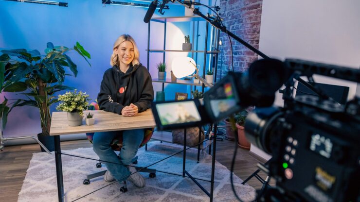 Snimanje video sadržaja u studiju s profesionalnom kamerom, rasvjetom i mikrofonom, dok djevojka sjedi za stolom i gleda prema objektivu.